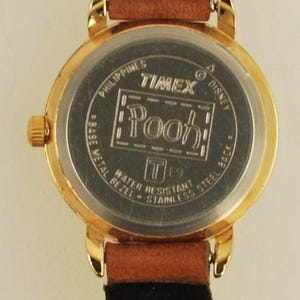 May include: A gold-toned Timex watch with a brown leather strap and black backing. The watch face features the word "Pooh" in a rectangular frame. The watch is water-resistant and has a stainless steel back.