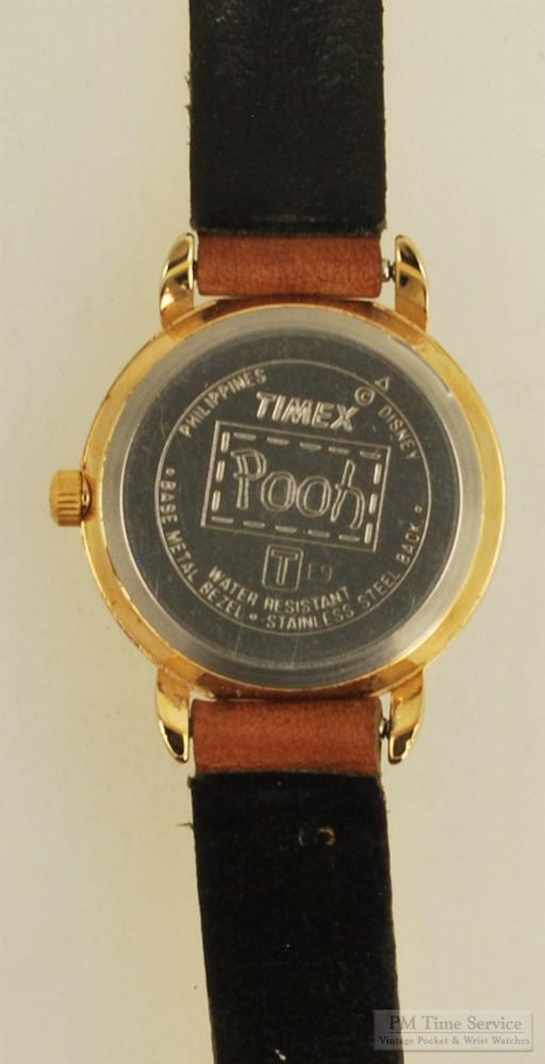May include: A gold-toned Timex watch with a brown leather strap and black backing. The watch face features the word "Pooh" in a rectangular frame. The watch is water-resistant and has a stainless steel back.