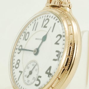 May include: A gold-toned pocket watch with a white face and black hands. The watch has a small second hand dial at the bottom. The watch face has black Roman numeral hour markers and black Arabic numeral minute markers. The watch has a gold-toned chain attached to the top.