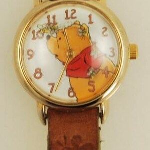May include: Gold-tone wristwatch featuring Winnie the Pooh on the face, with a brown leather band. The watch face has a white background with black numerals. The band is embossed with a floral pattern.
