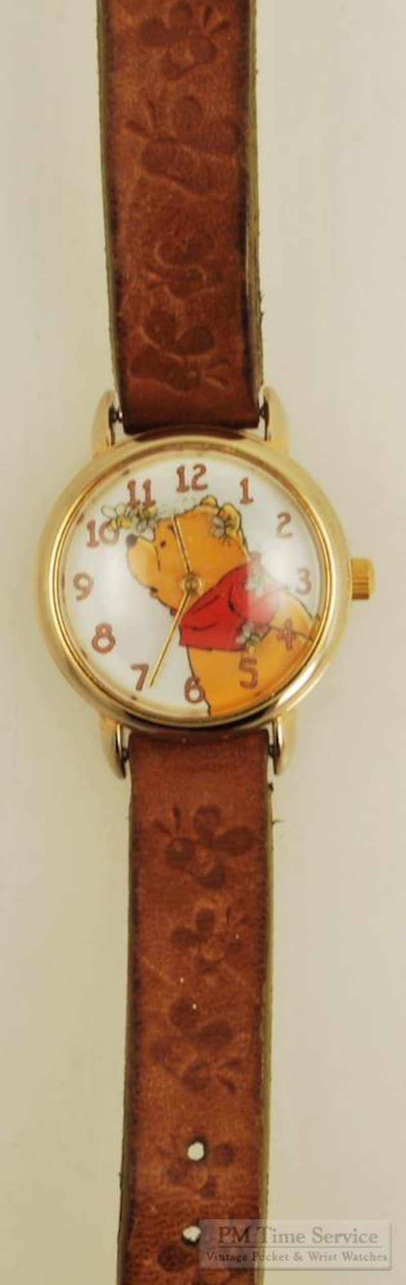 May include: Gold-tone wristwatch featuring Winnie the Pooh on the face, with a brown leather band. The watch face has a white background with black numerals. The band is embossed with a floral pattern.