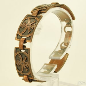 Op de afbeelding: Een vintage koperen armband met een geometrisch ontwerp. De armband heeft rechthoekige schakels met een centraal gestileerd ontwerp, verbonden door kleinere schakels. Het metaal heeft een warme, verouderde patina. De armband wordt op een transparante standaard getoond.