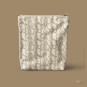 K&ouml;nnte beinhalten: Eine rechteckige Rei&szlig;verschlusstasche mit einem vertikalen abstrakten Muster in Creme-, Beige- und Metallic-Goldt&ouml;nen. Der Stoff hat eine strukturierte Optik mit einem sich wiederholenden organischen Design und einem silberfarbenen Metallrei&szlig;verschluss.