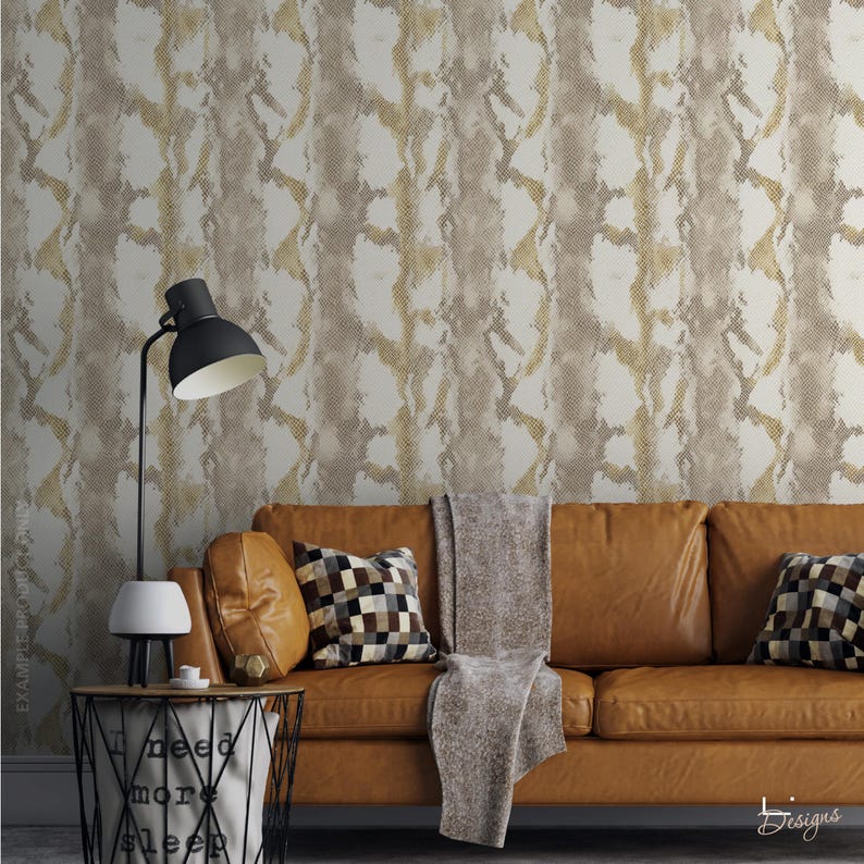 K&ouml;nnte beinhalten: Ein beiges Ledersofa mit einer grauen Decke und zwei karierten Kissen vor einer Wand mit wei&szlig;-goldener Schlangenhaut-Tapete. Eine schwarze Lampe steht auf einem Draht-Beistelltisch mit dem aufgedruckten Text I need more sleep.