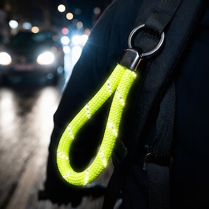 Puede incluir: Un llavero reflectante amarillo neón sujeto a una correa de mochila negra. El llavero está hecho de un material grueso y tejido con detalles blancos reflectantes. El fondo muestra una calle de la ciudad por la noche con faros de coche.
