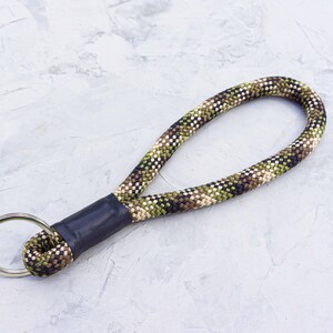 Peut inclure: Dragonne de porte-clés à motif camouflage avec un porte-clés en métal argenté. La sangle est faite d'une corde tissée dans des tons de vert, marron et beige, avec une bande noire reliant la corde au porte-clés.