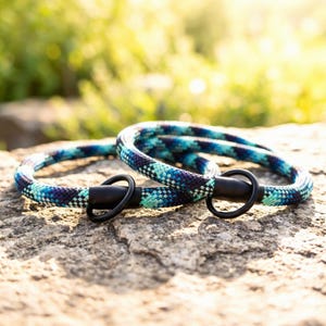 Op de afbeelding: Twee honden halsbanden gemaakt van een blauw, turquoise en paars touw met zwarte metalen hardware. De halsbanden liggen op een grijze stenen ondergrond met een vage groene achtergrond.