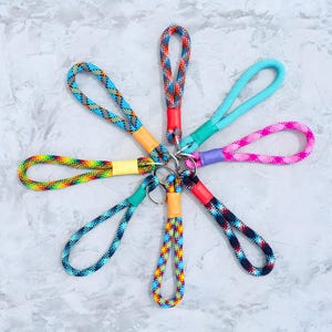 May include: A collection of colorful rope keychains arranged in a circular pattern. Each keychain features a loop of patterned rope in various colors, including blue, green, pink, and rainbow hues, connected to a metal ring with a colored band.