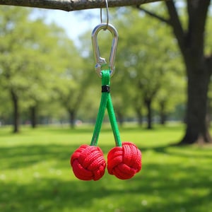Op de afbeelding: Een decoratieve sleutelhanger met twee rode, geknoopte ballen die op kersen lijken, opgehangen aan een groen koord en een zilveren karabijnhaak. De sleutelhanger hangt aan een tak tegen een vage groene achtergrond.