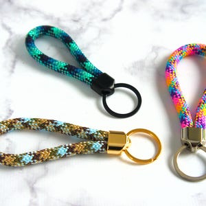 May include: Three keychains with looped wrist straps. One is multicolored, with pink, yellow, and blue. Another is green and blue. The third is brown, blue, and gold. Each has a metal ring for keys.