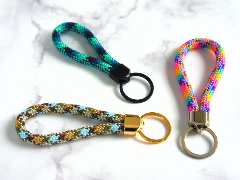 May include: Three keychains with looped wrist straps. One is multicolored, with pink, yellow, and blue. Another is green and blue. The third is brown, blue, and gold. Each has a metal ring for keys.