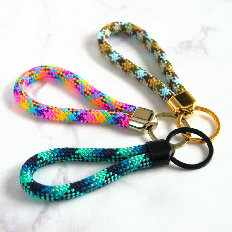 May include: Three colorful rope keychains. One is rainbow-colored, another is teal and navy, and the third is brown, tan, and blue. Each keychain has a metal clasp and a keyring. The keychains are on a white marble surface.