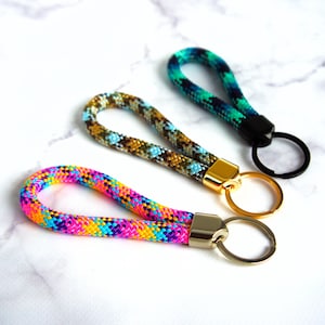 Pode incluir: Tr&ecirc;s chaveiros tecidos coloridos. Um &eacute; arco-&iacute;ris, outro tem um padr&atilde;o marrom, azul e dourado, e o terceiro &eacute; verde-azulado e verde. Cada chaveiro tem um fecho e um anel de metal.