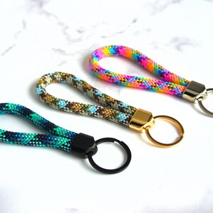 May include: Three colorful keychains with looped rope handles. One is teal and navy, another is brown and blue, and the third is rainbow-colored. Each has a metal clasp and keyring in gold, silver, or black.