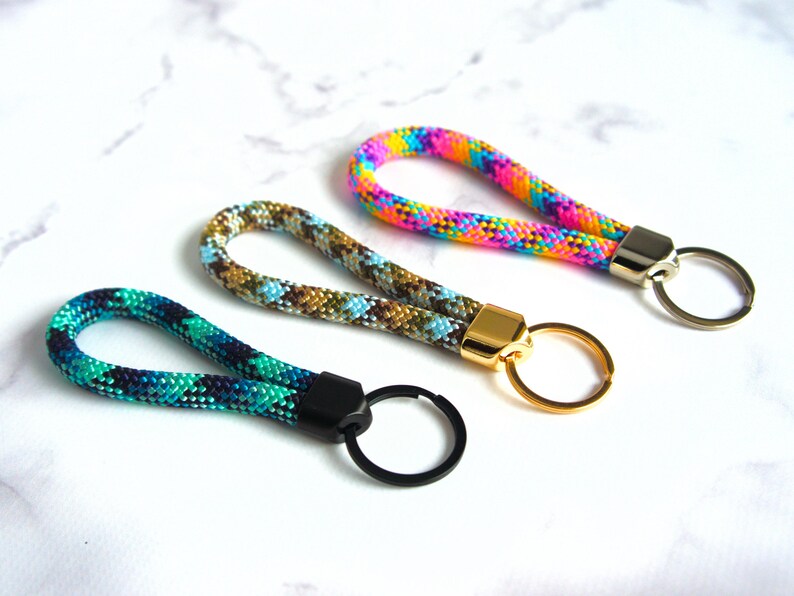 May include: Three colorful keychains with looped rope handles. One is teal and navy, another is brown and blue, and the third is rainbow-colored. Each has a metal clasp and keyring in gold, silver, or black.