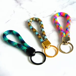 May include: Three keychains with woven rope straps in vibrant colors. One is teal and navy, another is brown and blue, and the third is rainbow-colored. Each keychain has a metal ring and a metal clasp in gold or black.