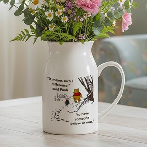Puede incluir: Jarra de cerámica blanca con asa, llena de un ramo de flores rosas, blancas y moradas. La jarra presenta una ilustración de Winnie the Pooh y la cita: "Marca tanta diferencia, dijo Pooh, tener a alguien que crea en ti."
