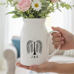 Puede incluir: Jarra de cerámica blanca con un diseño de árbol negro y las iniciales "J & S". La jarra contiene un ramo de rosas rosas, margaritas blancas y vegetación. Se ve un asa en el lado derecho.