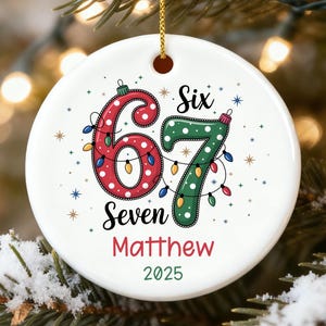 May include: A white ceramic Christmas ornament with the text "Six Seven Matthew 2025". The numbers 6 and 7 are decorated with polka dots and Christmas lights. The ornament is hanging from a gold string.