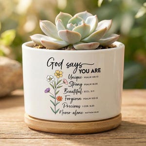 May include: White ceramic plant pot with a succulent. The pot features the text "God says YOU ARE" and floral illustrations. It rests on a round wooden base. The background is blurred with green foliage.