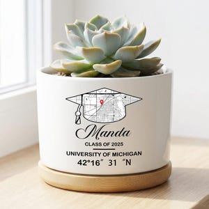 May include: A white ceramic planter with a succulent plant. The planter features a graduation cap design with a map, the name "Manda", and "University of Michigan". The planter sits on a wooden base.