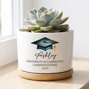 May include: A white ceramic planter with a succulent plant. The planter features a graduation cap design, the name "Ashley", and "University of Cambridge, Cambridgeshire 2025". The planter sits on a wooden base.