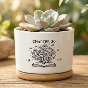 Peut inclure: Un pot de fleurs cylindrique en céramique blanche avec une base en bambou, contenant une plante succulente verte. L'avant présente un dessin au trait noir d'un livre ouvert avec des fleurs, des papillons, des étoiles et le texte "CHAPTER 30" et "19 96".