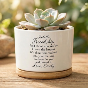 Może przedstawiać: Biała ceramiczna doniczka z sukulentem. Doniczka ma drewnianą podstawę i zawiera tekst "Isabella Friendship" oraz cytat o przyjaźni. Sukkulent ma jasnozielone liście.