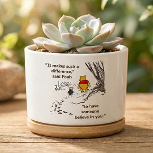 Puede incluir: Maceta de cerámica blanca con una planta suculenta. La maceta presenta una ilustración y una cita de Winnie the Pooh. La maceta se asienta sobre una base de madera. El texto dice: "Marca tal diferencia, dijo Pooh, tener a alguien que crea en ti."