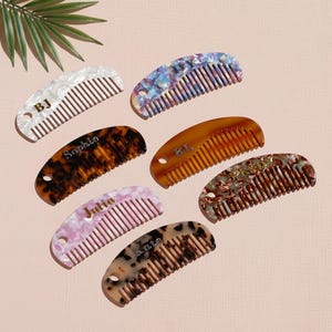 Puede incluir: Una variedad de peines personalizados en varios colores y patrones, incluyendo diseños blancos, azules, carey, rosas y moteados. Cada peine presenta un nombre o iniciales, como "BJ", "Sophie" y "Julia". Los peines están dispuestos sobre una superficie rosa claro.