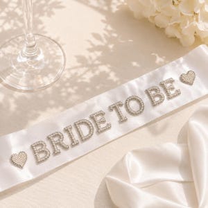 Puede incluir: Una banda de satén blanco con la frase "BRIDE TO BE" en letras plateadas brillantes. Dos adornos en forma de corazón con pedrería plateada en los extremos. Un accesorio nupcial.
