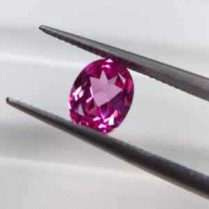 May include: A vibrant, oval-cut, pink gemstone is held by a pair of tweezers. The gemstone has a faceted surface, reflecting light. The background is a soft, neutral white.
