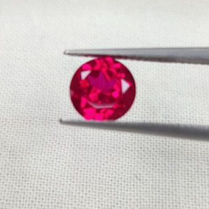 May include: A round, faceted, bright red gemstone is held by silver tweezers against a white background. The gemstone is a vibrant shade of red, with multiple facets catching the light. The tweezers are positioned to hold the gem securely.