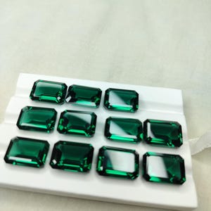 Puede incluir: Gemas verdes esmeralda de corte rectangular dispuestas sobre una superficie blanca. Las piedras son facetadas, reflejando la luz. Adecuadas para la creación de joyas o manualidades.