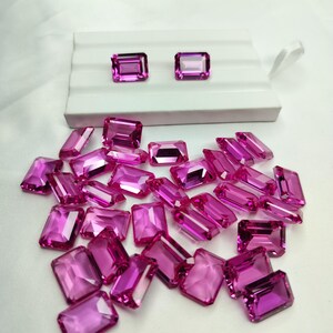 Puede incluir: Una colección de gemas rosas radiantes, de corte rectangular. Dos gemas se exhiben en una bandeja blanca, mientras que el resto están dispersas sobre una superficie blanca. Las gemas tienen una apariencia pulida y transparente, reflejando la luz.