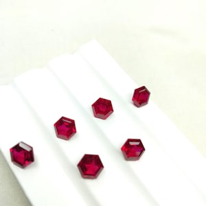 Peut inclure: Six pierres précieuses hexagonales, rouge foncé, sont disposées sur une surface blanche pliée. Les gemmes facettées ont un aspect poli, reflétant la lumière. L'arrangement suggère une présentation de pierres précieuses en vrac, peut-être pour la joaillerie ou la collection.