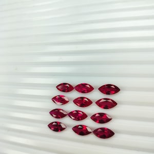 May include: Marquise-cut, deep red gemstones arranged in rows on a white, ridged surface. The gems have a pointed oval shape, with facets catching the light. The color is a rich, saturated red, suggesting ruby or garnet.