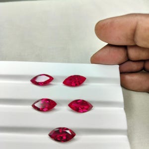 May include: Five marquise-cut, vibrant red gemstones are displayed on a white tray. The gems are arranged in two rows, with the bottom row containing a single gem. The gems are faceted, reflecting light.