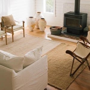May include: A living room scene featuring neutral-toned rugs, a white sofa, and wooden chairs. Three large, aged ceramic vases stand near a fireplace with a black stove. The room has a cozy, natural aesthetic.