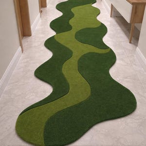 Può includere: Un tappeto verde ondulato in un corridoio, con una sezione centrale verde chiaro. Il design del tappeto crea un motivo fluido e organico. Il corridoio ha pareti e pavimenti di colore chiaro. Il tappeto misura circa 300 cm di lunghezza.