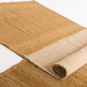 May include: A rectangular, woven jute rug in shades of tan and beige. The rug is partially rolled up, revealing a lighter beige underside. The texture of the rug is visible, with a natural, earthy aesthetic. The rug is suitable for home decor.