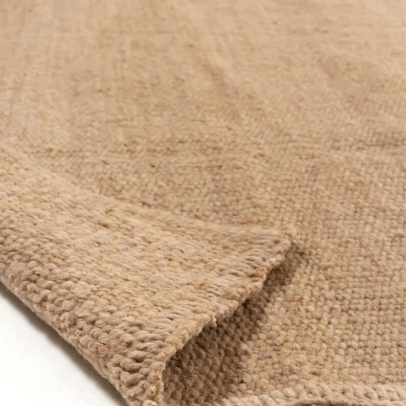 May include: A close-up of a beige woven rug with a textured surface. The rug has a folded edge, revealing the thickness and weave of the material. The color is a warm, neutral tone.