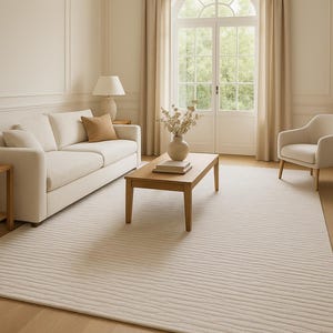 Peut inclure: Salon aux tons neutres avec un canapé et un fauteuil crème, une table basse en bois et un grand tapis texturé. La pièce est éclairée par une grande fenêtre cintrée et des rideaux beiges.