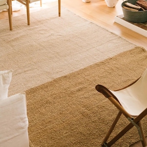 May include: A rectangular jute rug with a two-tone design. The top portion is a light beige, transitioning to a darker tan at the bottom. The rug is placed on a light wood floor, with a wooden chair and canvas chair visible.