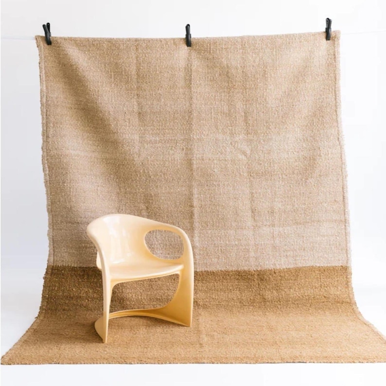 May include: A beige woven rug with a darker brown border is suspended from a white wall with black clips. A light beige chair with a curved design sits in front of the rug. The rug is made of a thick, textured material.