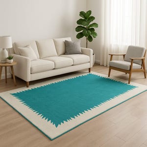 Puede incluir: Una alfombra rectangular con un centro turquesa y un borde color crema. El borde presenta un patrón geométrico repetitivo. La alfombra está colocada sobre un suelo de madera clara en un salón.