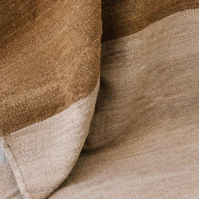 May include: Close-up of a woven rug with a textured surface. The rug features a color block design with shades of brown and beige. The fabric appears to be thick and durable, with a slight sheen.