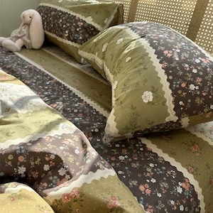May include: Bedding set featuring a floral pattern in shades of green, brown, and white. The set includes pillowcases and a duvet cover with a lace trim. A stuffed bunny is resting on the bed. The overall theme is a vintage, cottagecore aesthetic.
