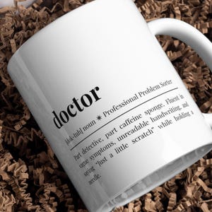 Könnte beinhalten: Weißer Keramikbecher mit dem Wort "doctor" in fetter schwarzer Schrift. Der Becher enthält eine Definition von "doctor" mit Sätzen wie "teilweise Koffeinschwamm" und "unleserliche Handschrift".