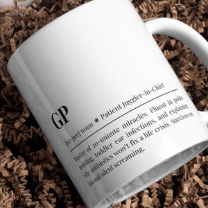 Puede incluir: Taza de cerámica blanca con las letras "GP" y una definición de "Patient Juggler-in-Chief". El texto describe las habilidades de un médico de cabecera, incluyendo el manejo de infecciones de oído en niños pequeños y la supervivencia con té. La taza tiene un asa curva.